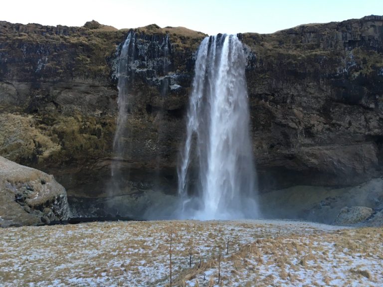 A tall waterfall cascading down a rocky cliff into a small pool, surrounded by grass.