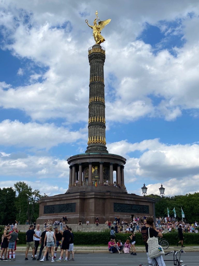 Victory Column Victory Column with golden statue, surrounded by people and greenery under a blue sky.