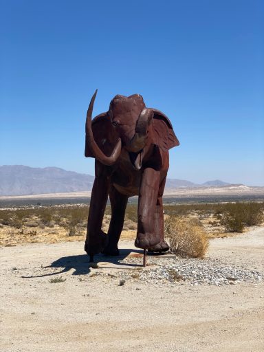 A rusty metal sculpture of an elephant standing in a desert landscape.