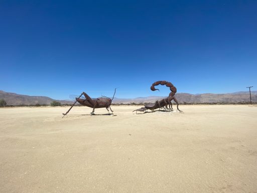 Metal sculptures of a grasshopper and a scorpion in a sandy desert landscape.