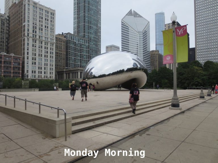 A reflective sculpture surrounded by tall buildings and people walking.