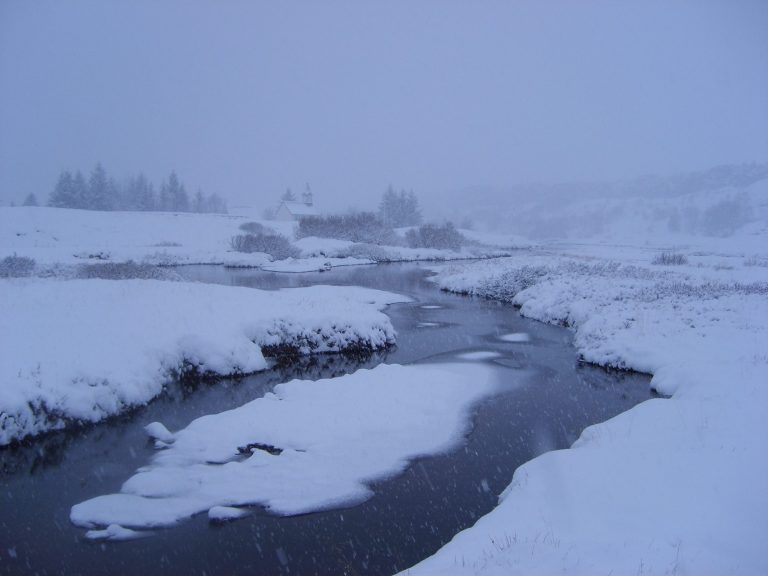 Snow-covered landscape with a river flowing through, surrounded by trees and fog.
