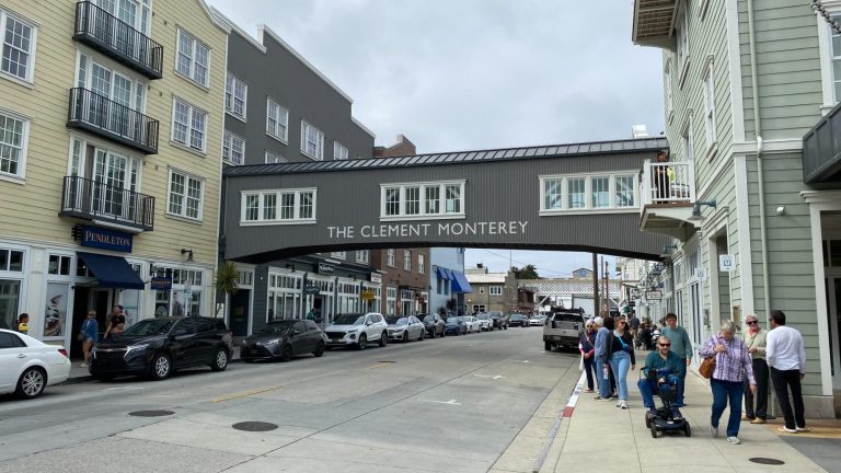 Street view of a pedestrian bridge, lined with parked cars and people.
