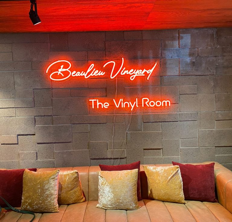 Neon sign reading "Beaulieu Vineyard The Vinyl Room" above a cushioned seating area.