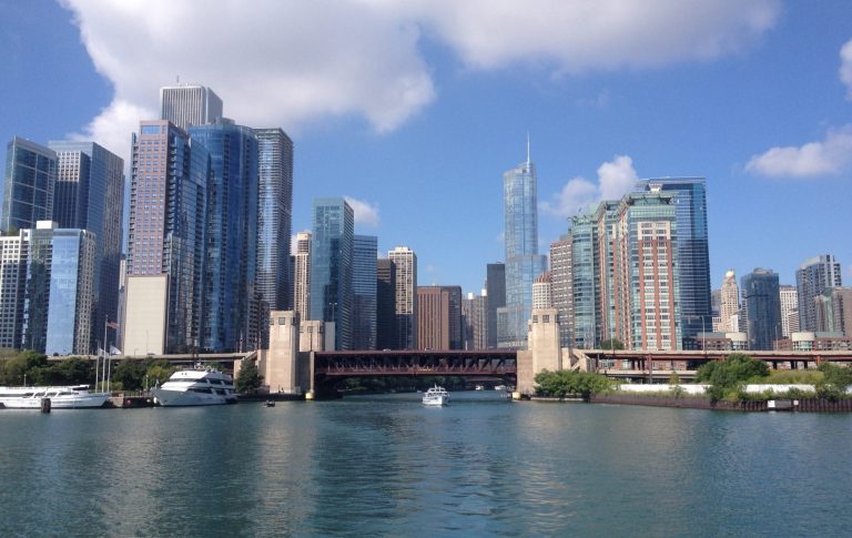 Skyline of tall buildings by a river on a clear day, with boats visible.