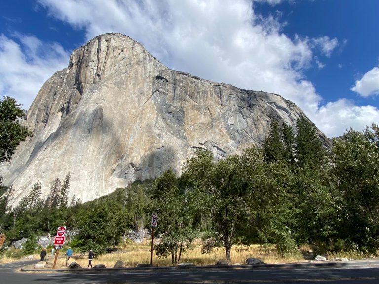 A large granite cliff surrounded by trees and blue sky with scattered clouds.