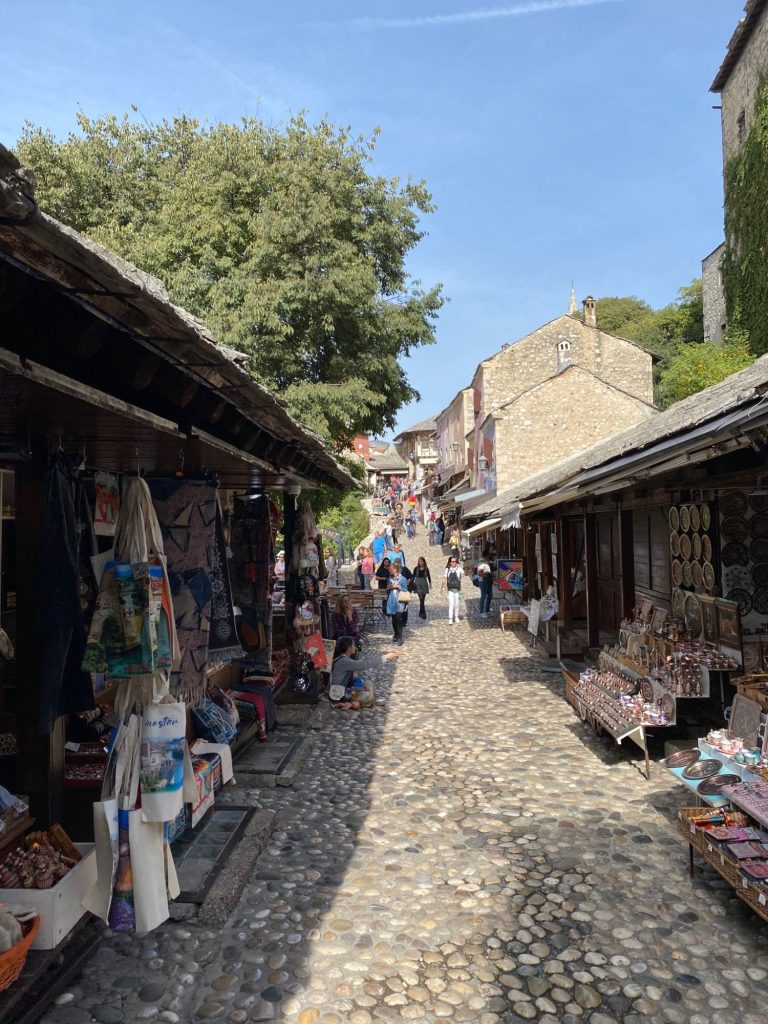 The Old Bazar, Mostar Narrow cobblestone street lined with shops and people, under a clear blue sky.