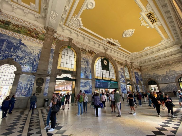 Interior of a grand train station with detailed tile murals and a bustling crowd.