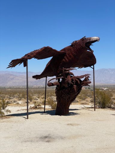 A large rusted sculpture of an eagle soaring above a hand, set against a desert backdrop.