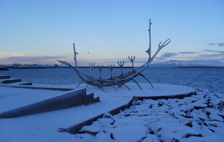 A snow-covered sculpture resembling a ship on a rocky shore, with a blue sky overhead.