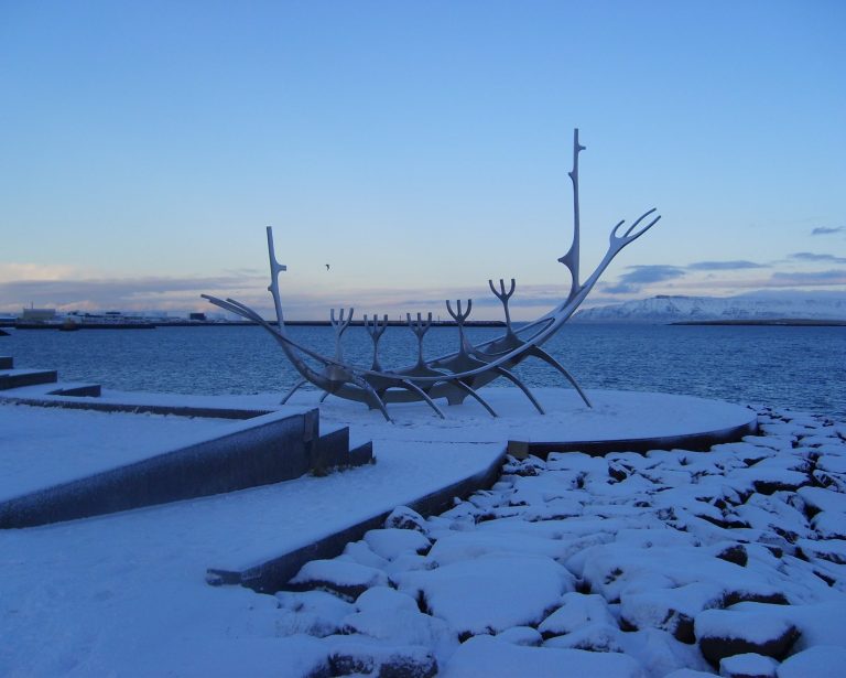 Sculpture resembling a boat on a snowy shore with a calm sea and cloudy blue sky.