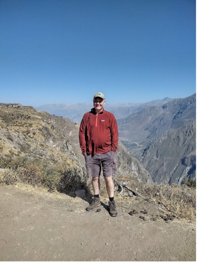 Man in a red jacket and shorts standing on a mountainous path with a blue sky.