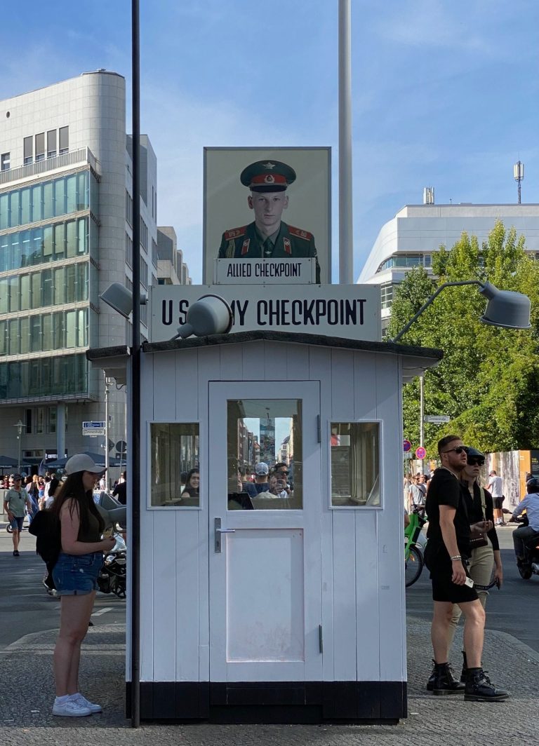 Checkpoint Charlie A small white booth with a sign featuring a military officer's portrait, near a busy street.