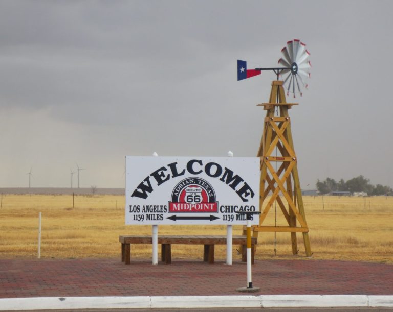 Welcome sign with windmill in a grassy landscape, featuring the Texas flag.
