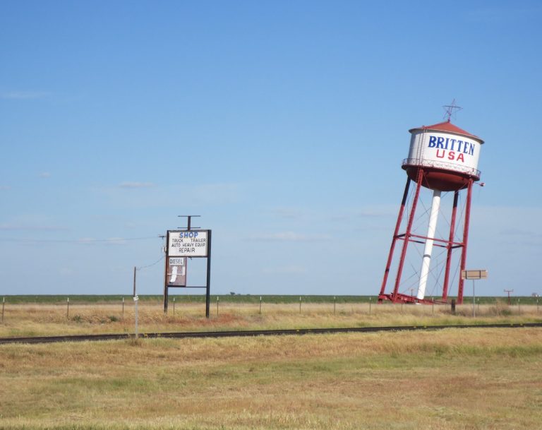 Water tower and a sign in a flat rural landscape under a clear blue sky.