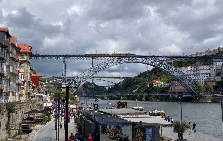 A bridge spans a river with cloudy skies and buildings along the bank.