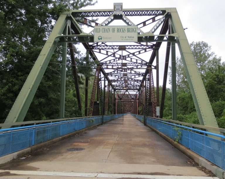 Green metal bridge with a blue railing, set against a backdrop of trees.