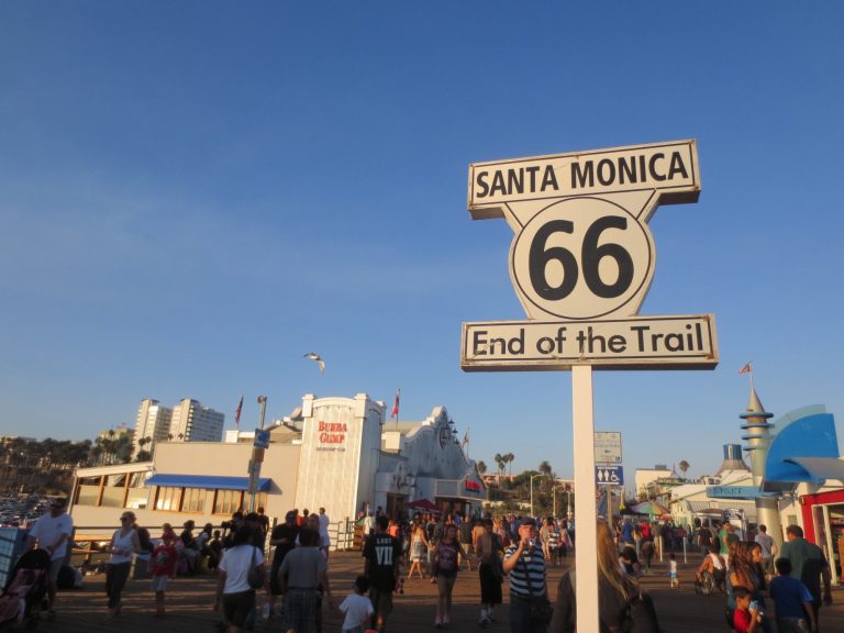 Signpost reading "Santa Monica 66 End of the Trail" with a bustling crowd in the background.