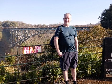 A man stands by a railing with a bridge in the background on a sunny day.