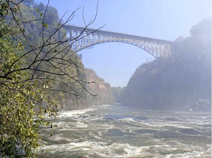 A tall arch bridge spans a misty river, surrounded by rocky cliffs and greenery.