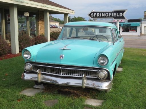 Vintage turquoise car parked in front of a shop, with a Springfield sign nearby.