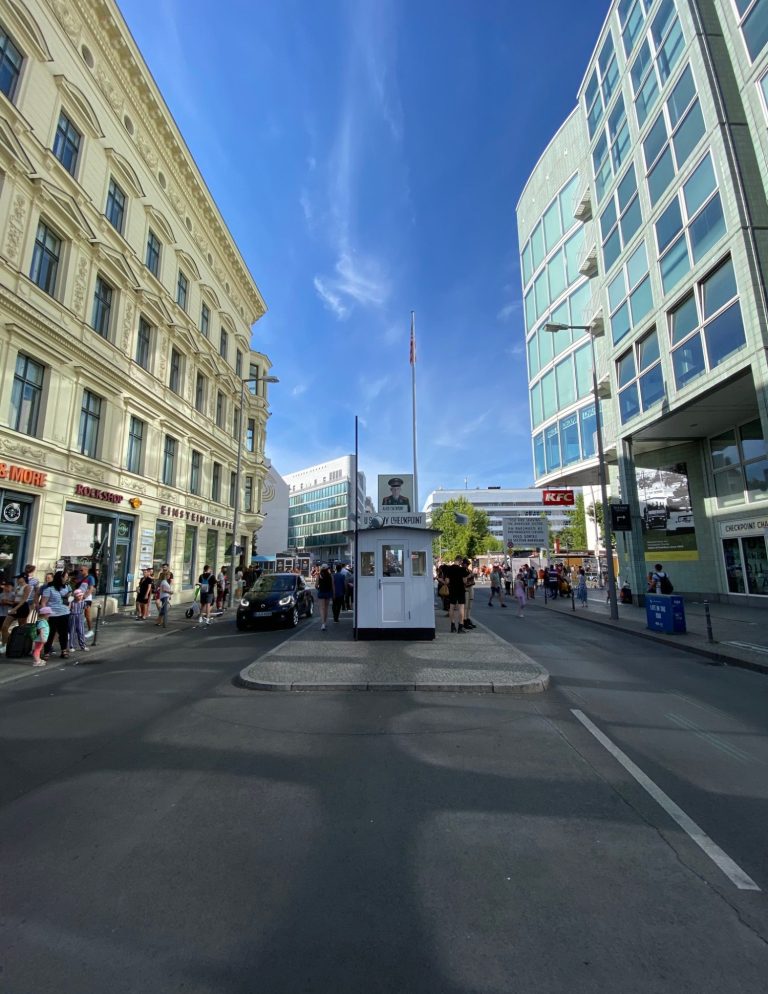 Checkpoint Charlie Busy street scene with people, buildings, and a white kiosk under a blue sky.