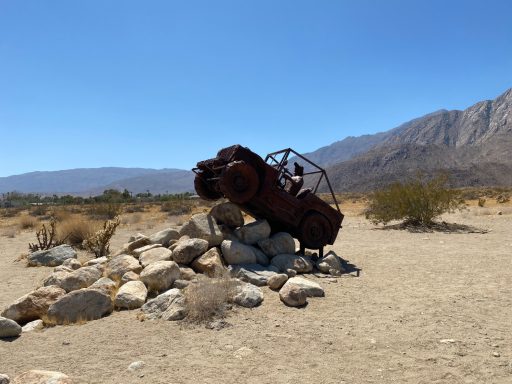 A rusted car precariously balanced on a pile of rocks in a desert landscape.