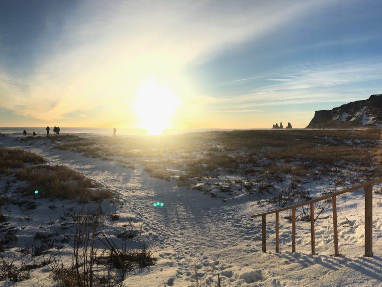 Sunset over a snowy landscape, with silhouettes of people and a wooden path on the right.