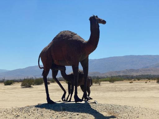 A large camel statue with a smaller camel beside it in a sandy desert landscape.