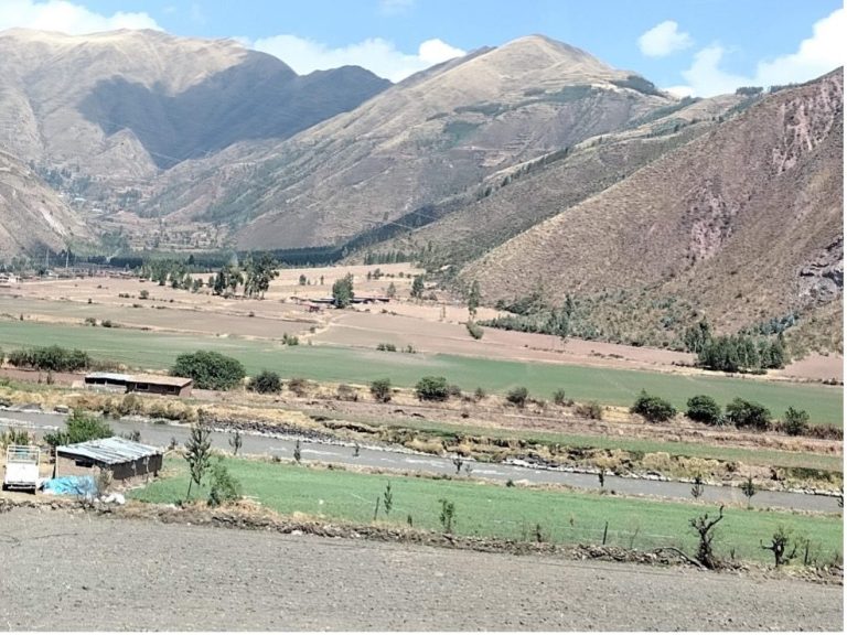 Lush green fields and a river set against a backdrop of mountains under a clear blue sky.