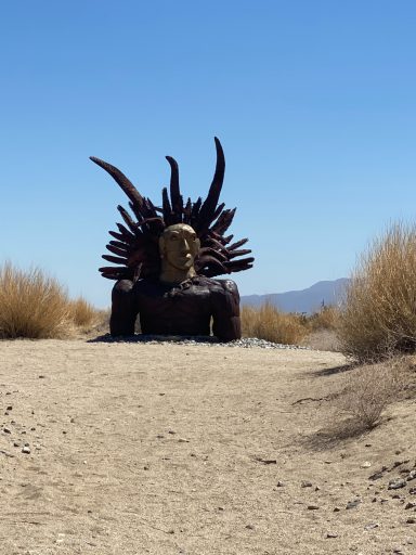A large, horned sculpture set in a sandy landscape with sparse vegetation.