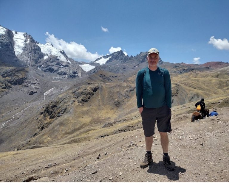 A person standing on a mountain trail with rocky terrain and snow-capped peaks in the background.