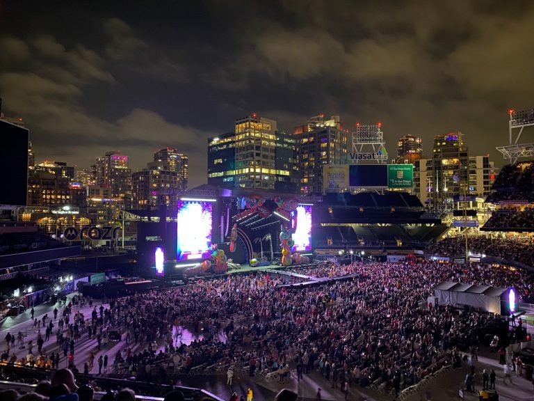 Crowd at an outdoor concert with city skyline and bright stage lights at night.