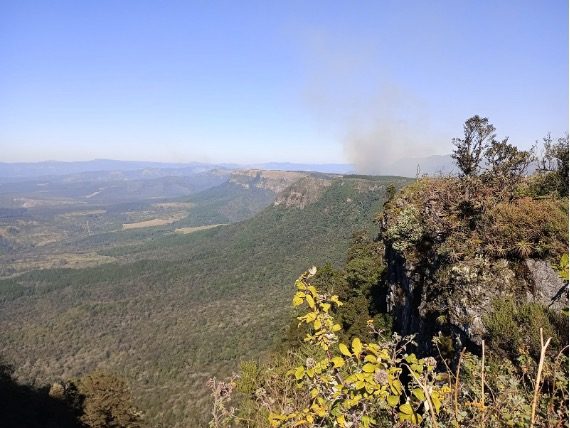 Vast mountainous landscape with distant smoke rising against a clear blue sky.