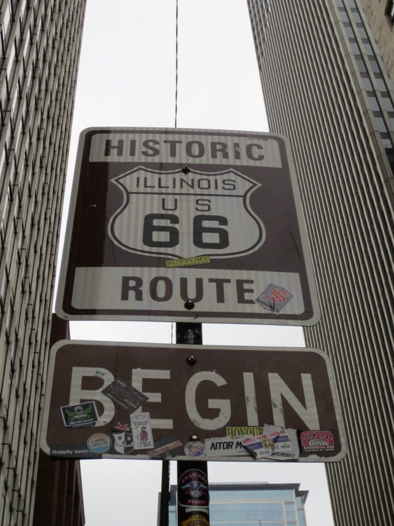 Sign marking the beginning of Historic Route 66 in Illinois, decorated with various stickers.