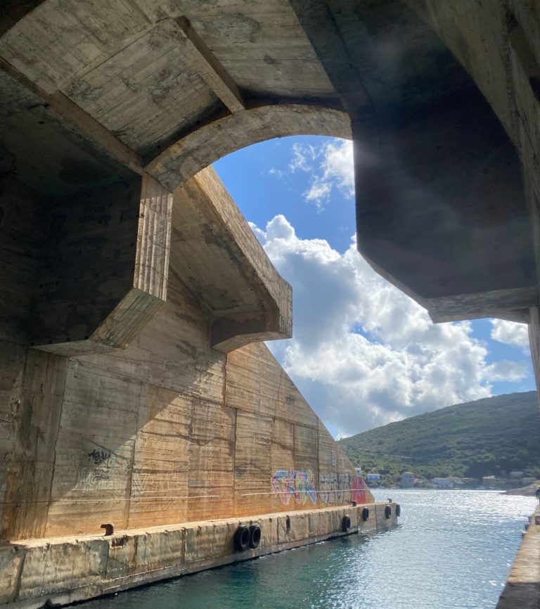 Yugoslavian Ship Hideout View through a concrete arch at a harbour, with blue sky and clouds reflected in the water.