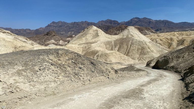 A rocky desert landscape with sand mounds and distant mountains under a clear blue sky.