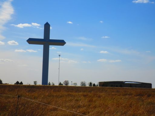 Large metal cross beside a building on a grassy landscape under a blue sky.