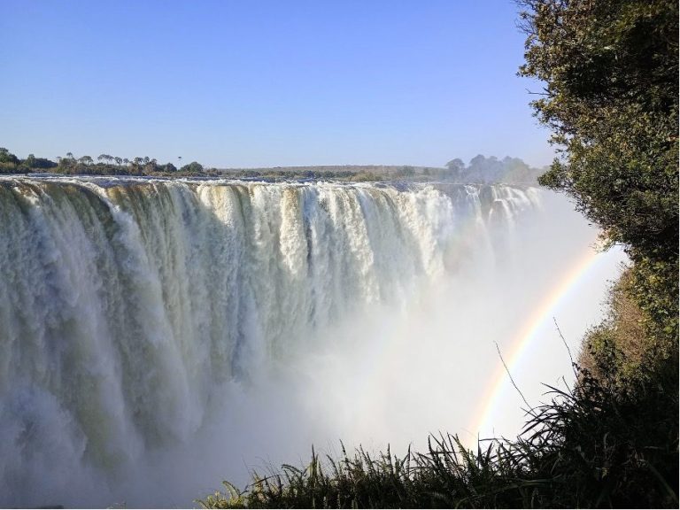 A view of a waterfall with a rainbow and lush greenery in the foreground.