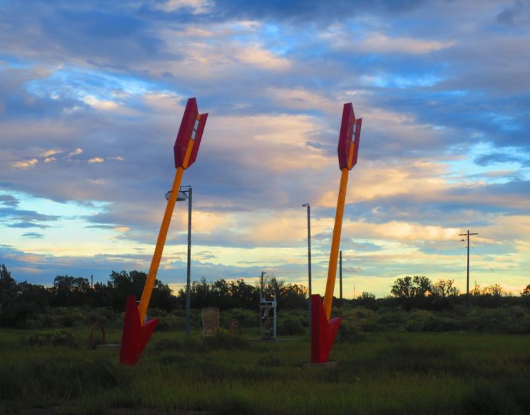 Two large red arrows point downwards in a grassy field under a cloudy sky.