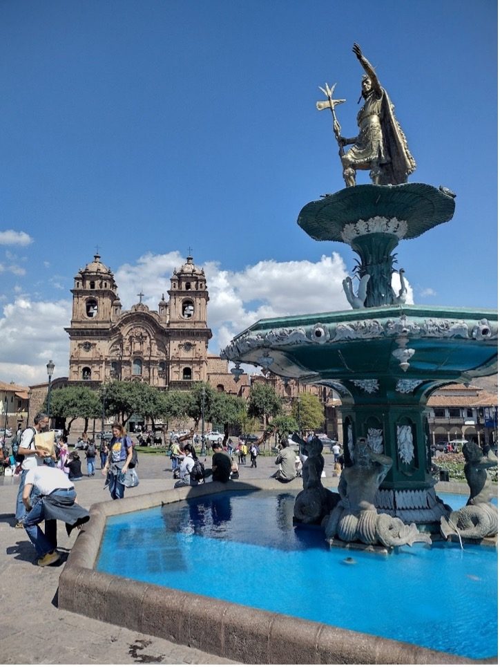 Fountain in a plaza with a church in the background and people gathered around.