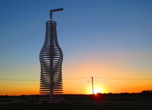 A large bottle sculpture with a straw against a sunset backdrop.