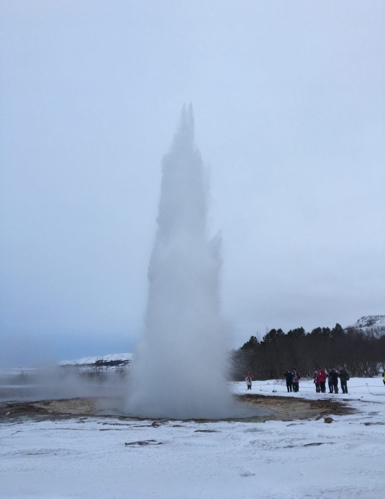 Geyser erupting with water and steam against a cloudy sky, surrounded by snowy ground.