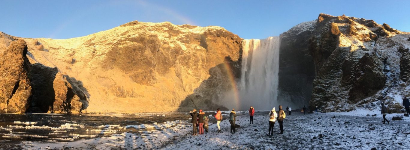 Panoramic view of a waterfall with people near the base, surrounded by snowy landscape.