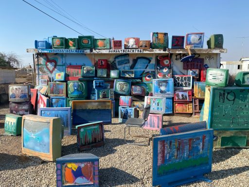 Colourful painted televisions and art pieces arranged outdoors against a blue sky.