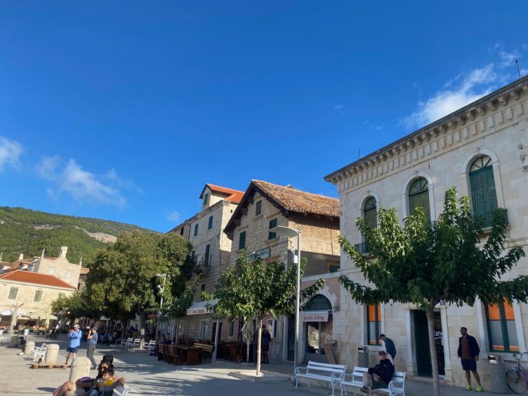 Komiza Town Historic buildings with green trees, blue sky, and people in a lively town square.